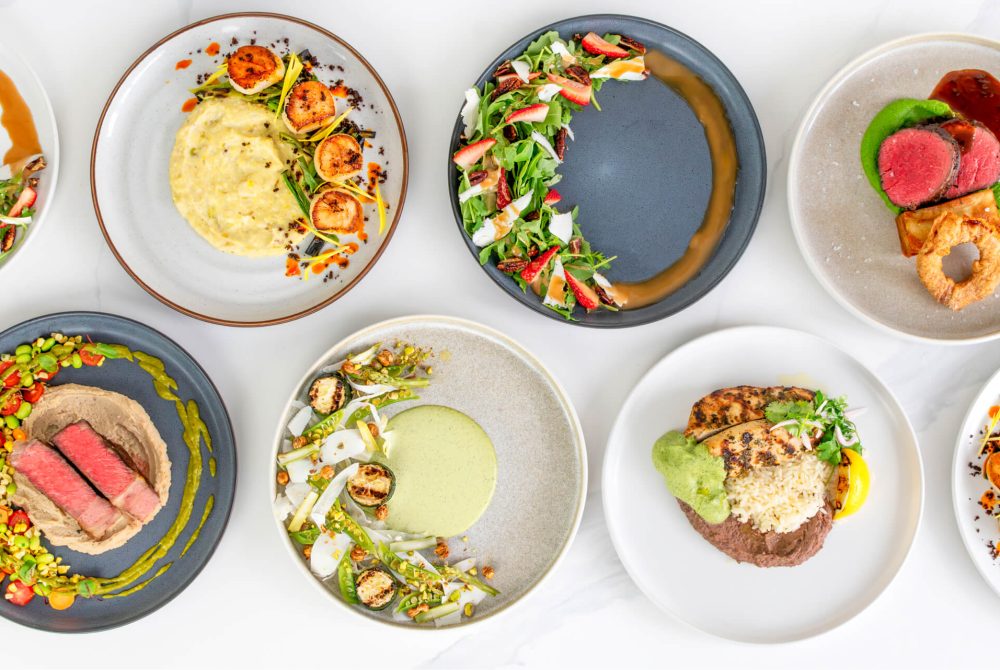 Overhead view of assorted plated dinner dishes by Palmer’s Catering & Events featuring seared scallops, steak, grilled chicken, fresh seasonal salads, and elegant vegetable sides.